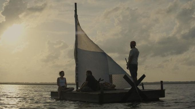 Still from the film The Peanut Butter Falcon. Two men and a woman are on a makeshift sailing boat in a body of water. The sun is setting.