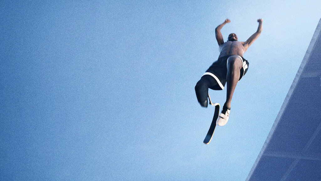 Still from the documentary Rising Phoenix. A from the ground shot of a Black man with one leg and a blade jumping in the air. The sky is a vast blue.